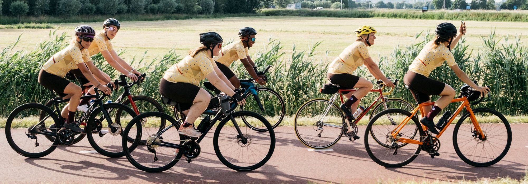 Five cyclists ride in a group along a paved path in an outdoor rural setting, wearing matching yellow jerseys and black shorts. Trees and grass are visible in the background, with one cyclist riding an orange bicycle.